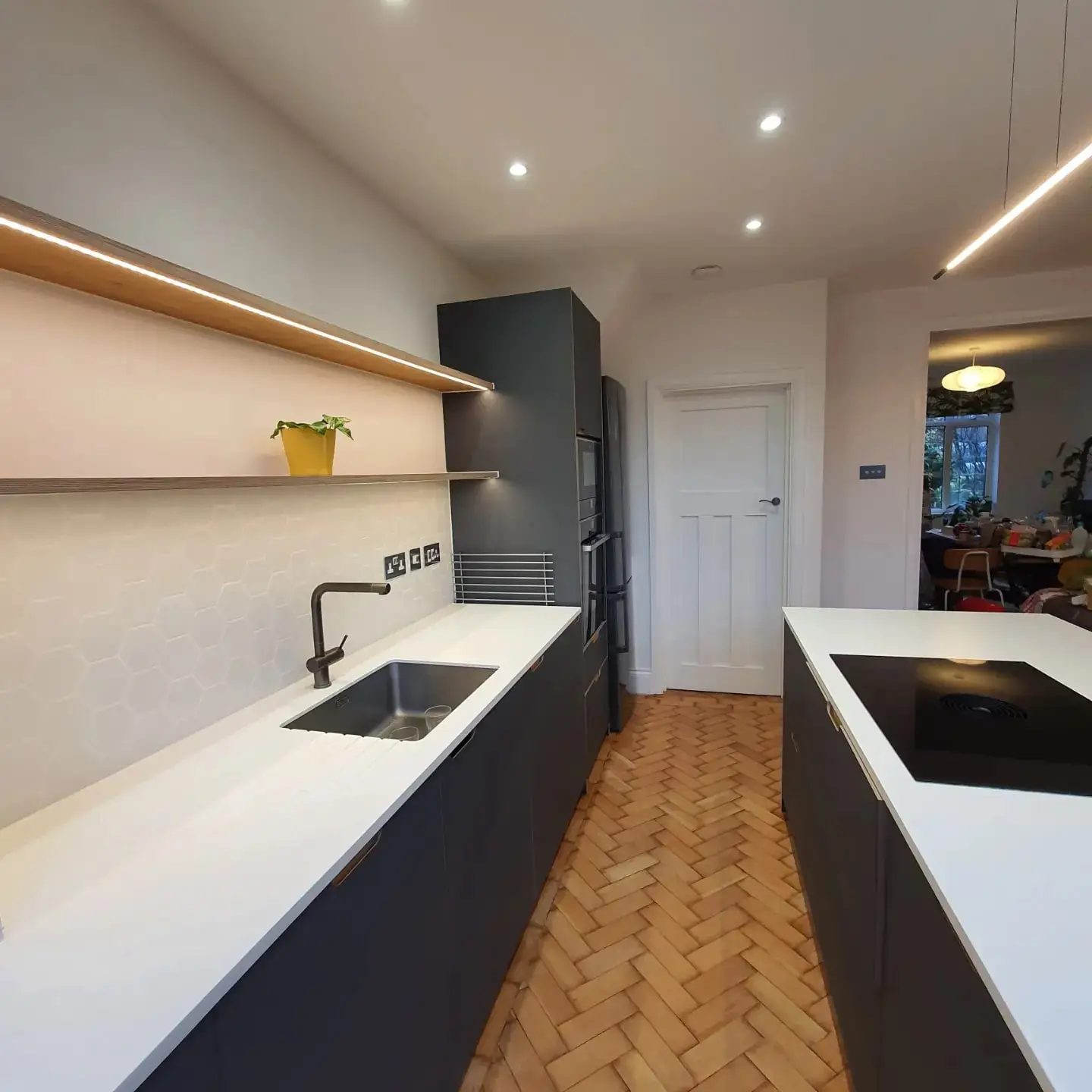 The new kitchen run in the extension: dark units, a hexagon tile splashback, lit oak shelves and a parquet floor